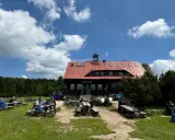 Chatka górzystów z czerwonym dachem, otoczona lasem. Ludzie odpoczywają przy stołach, obok stoją rowery.