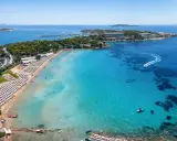 Traumhafter Blick auf das athen meer mit einer belebten Strandpromenade, Liegestühlen und dem glitzernden, türkisfarbenen Wasser, auf dem Boote fahren.