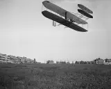 Das Flugzeug der Gebrüder Wright fliegt über ein Feld. Im Hintergrund sind Gebäude zu sehen.