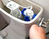 Mécanisme de chasse d'eau qui coule, avec un flotteur bleu et blanc visible. Une main ajuste le robinet d'arrivée d'eau.