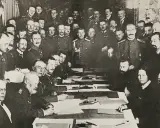 Delegierte unterzeichnen den Frieden von Brest-Litowsk. Männer in Uniformen und Anzügen sitzen an einem Tisch, umgeben von weiteren Personen.