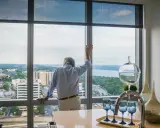 Hombre mira por ventana de aluminio, pensando cuánto pesa una ventana de aluminio. Vista urbana y río.