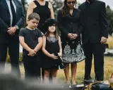 Dziecko na pogrzebie, w czarnej sukience i z misiem, stoi obok chłopca. Rodzina w żałobie, być może wierzą w przesądy związane z obecnością dzieci na ceremonii.