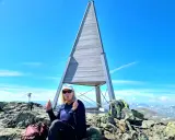 En kvinna sitter på toppen efter att ha lyckats bestiga Helags. Hon ger tummen upp vid den ikoniska träpyramiden.