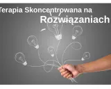 Dłoń trzymająca pęk żarówek narysowanych kredą na szarym tle.