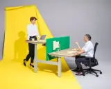Femme en position debout statique, souriante, devant un bureau. Homme assis à un bureau, discutant. Fond jaune et gris.