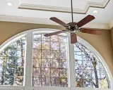 Ventilador de techo de madera oscura sobre una ventana arqueada con vista a árboles otoñales.