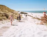 Familj på väg längs en träspång över vit sand i Danmark. En rödvit fyr och bunkrar syns i bakgrunden.