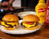 Två saftiga hamburgare med smält ost, pickles och lök på en tallrik. Bredvid står flaskor med senap och ketchup.