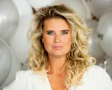 Blond kvinna med blå ögon och lockigt hår, omgiven av silverfärgade ballonger. Hon ser ut att vara redo för festliga udda veckor.