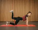 Femme effectuant un exercice culotte de cheval avec un élastique, sur un tapis de gym, devant un mur en bois.