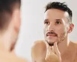 Mężczyzna w lustrze zastanawia się, jak zagęścić brodę na policzkach. Delikatnie dotyka zarostu, szukając rozwiązania.