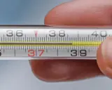 Zbliżenie na termometr rtęciowy, gdzie słupek rtęci wskazuje 37.1 temperatura. Dłoń trzyma termometr.