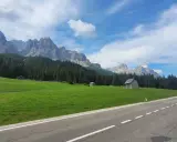 Strada panoramica verso il passo Monte Croce, con prati verdi, boschi di conifere e imponenti vette rocciose sotto un cielo azzurro con nuvole bianche.