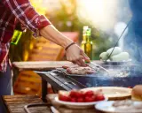Hombre asa salchichas envueltas en tocino, ideal para saber qué llevar a una barbacoa para sorprender. Hay cervezas y tomates en la mesa.