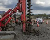 Máquina perforadora roja en obra, extrayendo tierra y formando columnas de grava. Conos naranjas y equipo de iluminación al fondo.