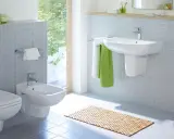 Nowoczesna łazienka z bidetem, który służy do higieny intymnej. Obok wisząca miska WC, umywalka i półki z ręcznikami.