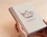Manos sostienen un libro con tapa de tela, con un diseño de tetera y el texto "La Posada del Té". Una idea para como encuadernar un trabajo de forma original.