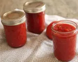 Trois bocaux de ketchup maison, un rouge vif, reposent sur une serviette à motifs. Un tomate entière est visible.