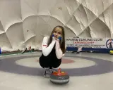 Młoda sportsmenka na lodzie, gotowa do gry w curling. W tle baner "Gdańsk Curling Club Zapraszamy".
