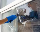 Ręce w niebieskich rękawiczkach myją okno w słońcu, używając ściągaczki i gąbki. Piana spływa po szybie.