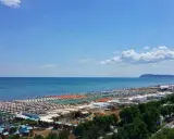 Ampia spiaggia di Riccione con file ordinate di ombrelloni e lettini, bagnanti che si godono il sole e l'acqua cristallina del mare Adriatico sotto un cielo azzurro.