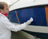 Un homme en combinaison blanche effectue la mise en cire d'un bateau bleu, protégeant la coque pour l'hiver.
