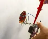 Ręce montują włącznik światła, podłączając przewody. Widać instalację elektryczną i narzędzie.