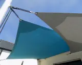 Comment installer un voile d'ombrage sur terrasse : deux voiles, bleu et gris, fixés par des cordages et des poulies à un mât et à la maison.
