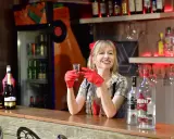 Uśmiechnięta barmanka w czerwonych rękawiczkach trzyma kieliszek. Na ladzie stoi duża butelka wódki, idealna na pół litra na dwóch.