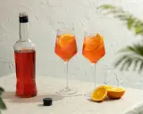 Dwa orzeźwiające Aperol Spritz z plasterkami pomarańczy, butelka Aperolu i połówki pomarańczy na lnianym obrusie.