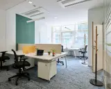 Un bureau chaleureux avec des postes de travail modernes, des cloisons acoustiques et un tapis bleu doux. L'espace est lumineux grâce aux grandes fenêtres.