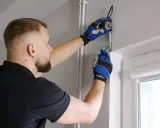 Mężczyzna w rękawiczkach mierzy taśmą i zaznacza ołówkiem miejsce do mocowania karnisza do sufitu.