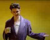 Janusz Walczuk w fioletowym szlafroku, z naszyjnikami i kubkiem z napisem "Wierzę w Ciebie!".