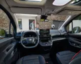 Interior de la nueva Citroën Berlingo, con pantalla táctil, volante moderno y asientos cómodos. Afuera, una pareja se acerca a un punto de carga.