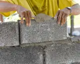 Budowa fundamentu z bloczków betonowych. Ręce murarza układają kolejny bloczek, używając kielni z zaprawą.