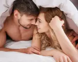 Para zbliża się do siebie pod kołdrą, ich twarze są blisko, sugerując intymność i potencjalny orgazm u kobiet.