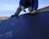 Un ouvrier en bleu, ganté, utilise un outil pour la pose d'un pare pluie sur un toit. Le ciel est bleu avec quelques nuages.