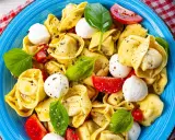 Ein bunter Tortellini Salat mit Mozzarella-Kugeln, Tomatenstücken und frischem Basilikum, serviert auf einem blauen Teller.