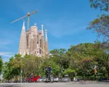 Sagrada Familia w Barcelonie, wciąż w budowie, góruje nad zielonym parkiem. Na ulicy widać rowerzystę i samochody.