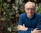 David Grossman, con occhiali blu e maglione scuro, sorride con le braccia incrociate su sfondo verde.