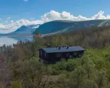 Mörkbrun byggnad vid en sjö, omgiven av björkskog och med fjäll i bakgrunden. En idyllisk plats för ritsem camping.