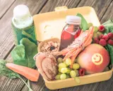 Zdrowa dieta 3 latka: żółty lunchbox z jabłkiem, malinami, winogronami, marchewką, placuszkiem i sokami.