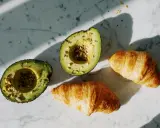 Dwa połówki awokado z nasionami i dwa chrupiące croissanty na marmurowym blacie. Idealne składniki na pyszne awokado przepisy.