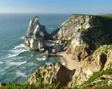 Steile Felsklippen umgeben einen einsamen sintra strand. Das türkisfarbene Meer bricht sich an den Felsen.