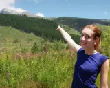 Uśmiechnięta dziewczyna wskazuje na górski krajobraz. W tle las i szczyty, a na pierwszym planie kwitnące chabry. To piękny krywań szlak.