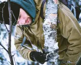 Mężczyzna w kurtce camo pochyla się nad drzewem w zimowym lesie.