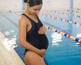 Kobieta w ciąży w czarnym stroju kąpielowym stoi przy basenie, delikatnie dotykając brzucha.