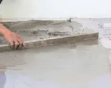 Ręka wyrównuje świeżą betonową wylewkę na ziemi za pomocą łaty. Kluczowe etapy, jak zrobić wylewkę betonową na ziemi.