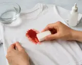 Manos aplicando un producto con una esponja para quitar manchas de sangre secas de una camiseta blanca.